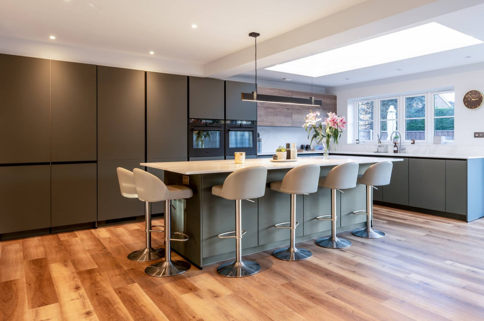 Photo of a large contemporary u-shaped eat-in kitchen in Hertfordshire with a single-bowl sink, flat-panel cabinets, green cabinets, quartzite benchtops, black appliances, medium hardwood floors, with island, brown floor and white benchtop.
