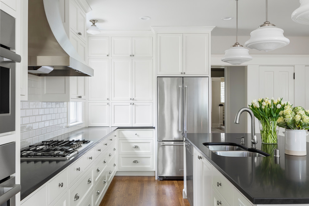 Example of a large transitional l-shaped medium tone wood floor and brown floor enclosed kitchen design in Minneapolis with an undermount sink, shaker cabinets, white cabinets, granite countertops, white backsplash, ceramic backsplash, stainless steel appliances, an island and black countertops