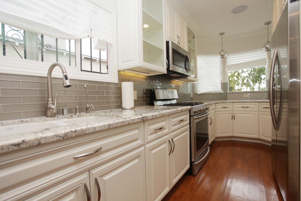Photo of a small traditional galley separate kitchen in Los Angeles with an undermount sink, raised-panel cabinets, white cabinets, granite benchtops, glass tile splashback, stainless steel appliances, dark hardwood floors, a peninsula, grey splashback and brown floor.