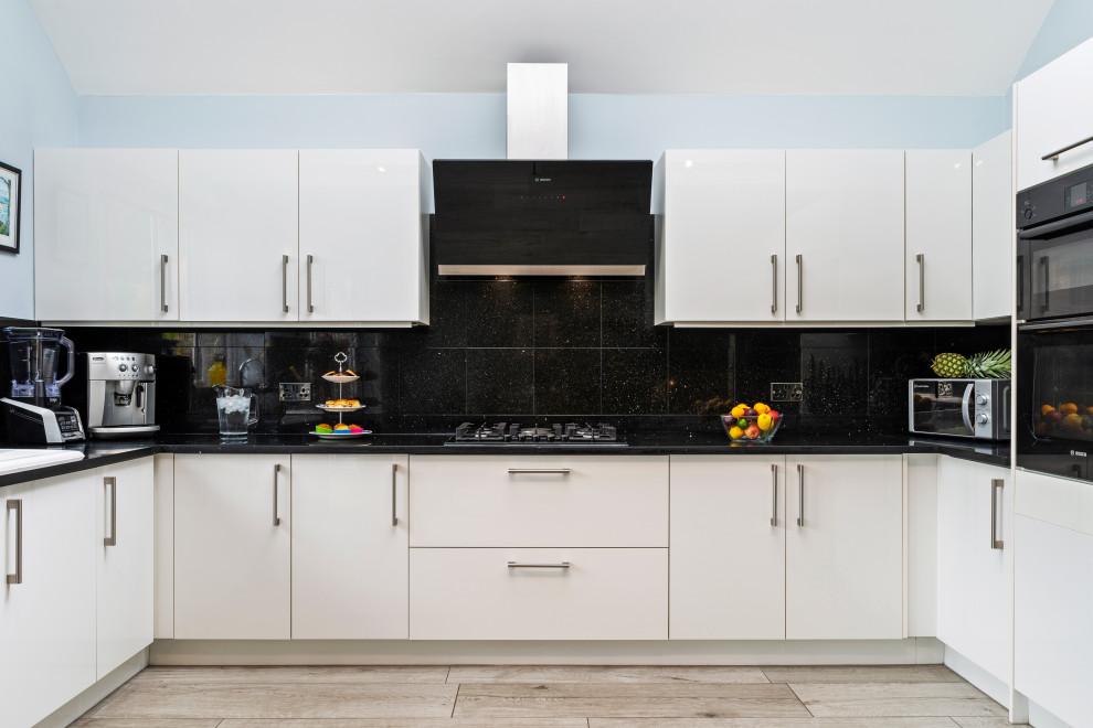 Inspiration for a mid-sized contemporary l-shaped open plan kitchen in Surrey with a drop-in sink, flat-panel cabinets, white cabinets, quartzite benchtops, black splashback, engineered quartz splashback, black appliances, porcelain floors, grey floor and black benchtop.