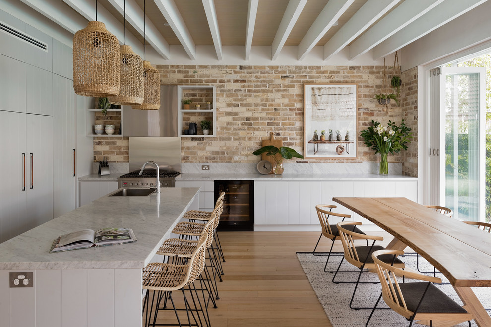 Inspiration for a beach style l-shaped eat-in kitchen in Sydney with a drop-in sink, flat-panel cabinets, white cabinets, brick splashback, stainless steel appliances, light hardwood floors and with island.