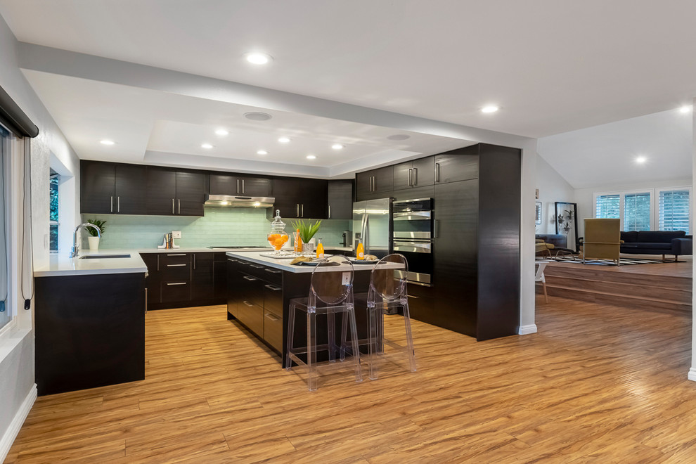Inspiration for a large contemporary u-shaped open plan kitchen in Los Angeles with an undermount sink, flat-panel cabinets, black cabinets, quartzite benchtops, grey splashback, glass tile splashback, stainless steel appliances, light hardwood floors, with island, brown floor and white benchtop.
