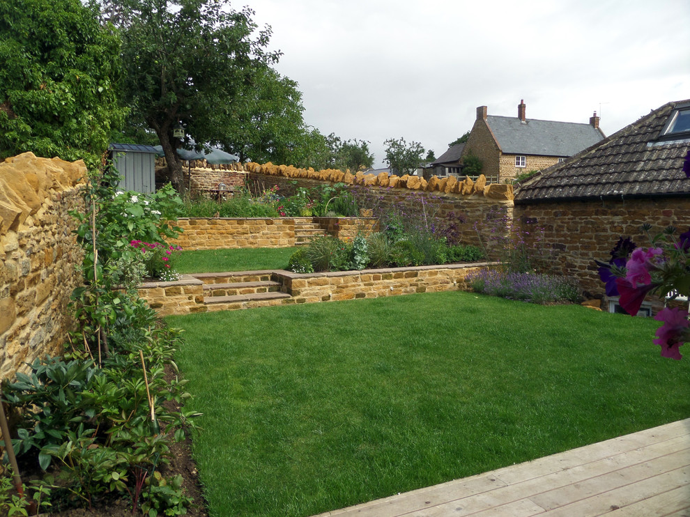This is an example of a mid-sized country sloped full sun garden for summer in Hertfordshire with a retaining wall and natural stone pavers.