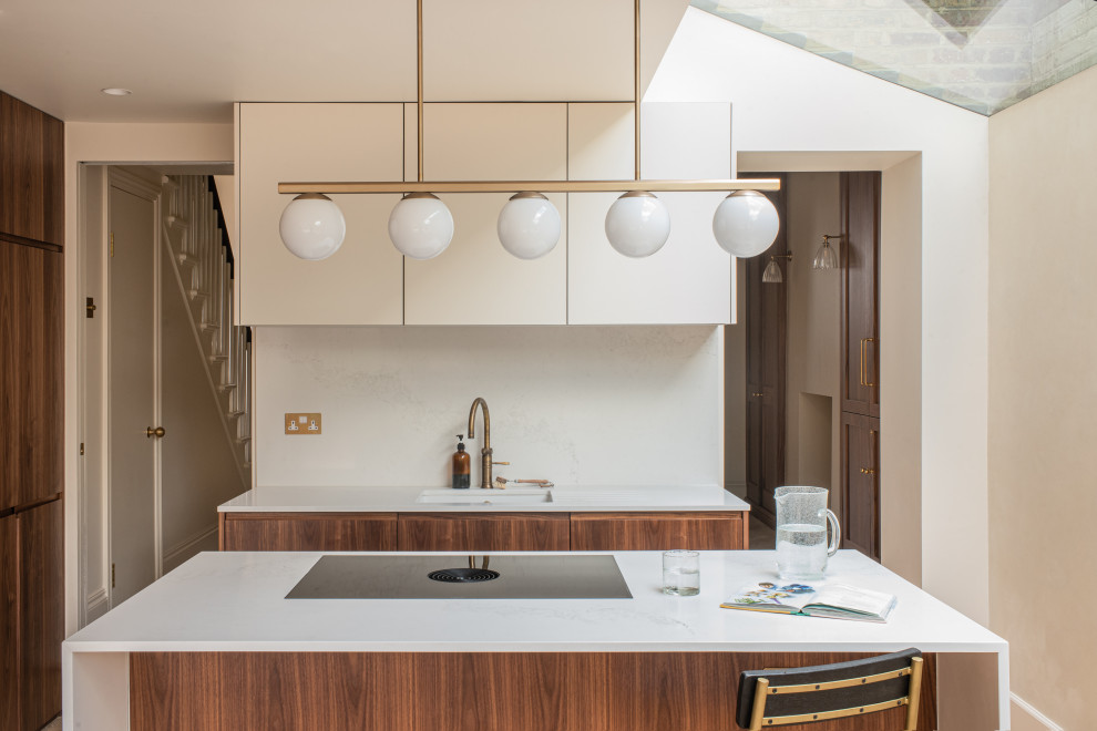 Inspiration for a mid-sized midcentury galley open plan kitchen in London with an integrated sink, flat-panel cabinets, dark wood cabinets, solid surface benchtops, beige splashback, black appliances, light hardwood floors, with island and beige benchtop.