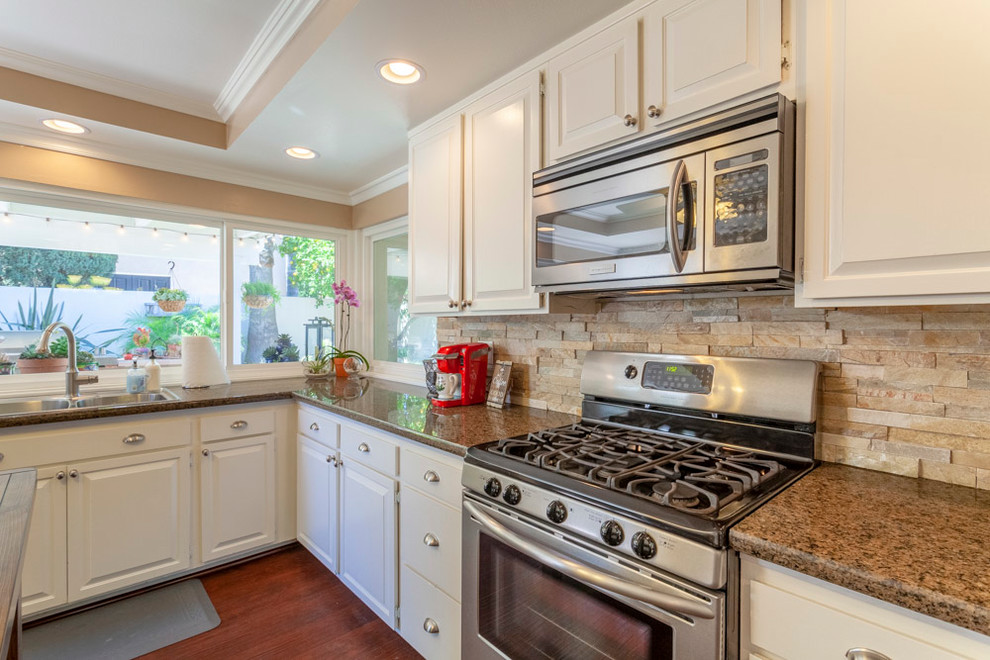 Inspiration for a mid-sized traditional u-shaped kitchen in Los Angeles with a drop-in sink, raised-panel cabinets, white cabinets, granite benchtops, white splashback, stone tile splashback, stainless steel appliances, medium hardwood floors, with island, brown floor and brown benchtop.
