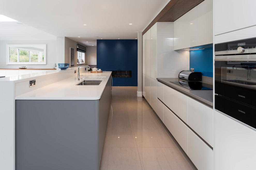 Expansive contemporary single-wall eat-in kitchen in Hertfordshire with a double-bowl sink, flat-panel cabinets, white cabinets, solid surface benchtops, blue splashback, glass sheet splashback, panelled appliances, porcelain floors, with island, grey floor and white benchtop.