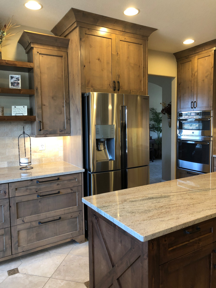 This is an example of a large country u-shaped eat-in kitchen in Denver with an undermount sink, shaker cabinets, distressed cabinets, granite benchtops, beige splashback, travertine splashback, stainless steel appliances, travertine floors, with island, beige floor and brown benchtop.