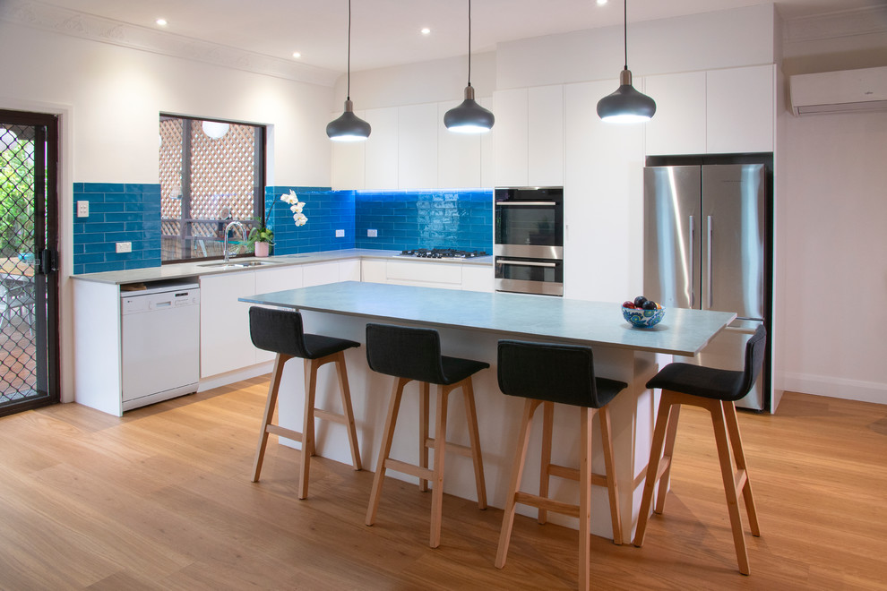 Mid-sized contemporary l-shaped open plan kitchen in Newcastle - Maitland with an undermount sink, flat-panel cabinets, white cabinets, quartz benchtops, blue splashback, subway tile splashback, stainless steel appliances, light hardwood floors, with island and grey benchtop.