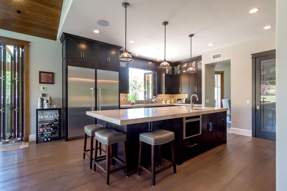Photo of an expansive arts and crafts u-shaped open plan kitchen in Orange County with a triple-bowl sink, recessed-panel cabinets, dark wood cabinets, quartzite benchtops, grey splashback, glass tile splashback, stainless steel appliances, medium hardwood floors, with island and grey floor.