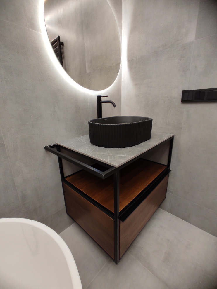 Пример оригинального дизайна: серо-белая ванная комната среднего размера в современном стиле с фасадами цвета дерева среднего тона, отдельно стоящей ванной, душем без бортиков, инсталляцией, серой плиткой, керамогранитной плиткой, серыми стенами, бетонным полом, душевой кабиной, подвесной раковиной, столешницей из гранита, серым полом, шторкой для ванной, серой столешницей, зеркалом с подсветкой, тумбой под одну раковину и подвесной тумбой