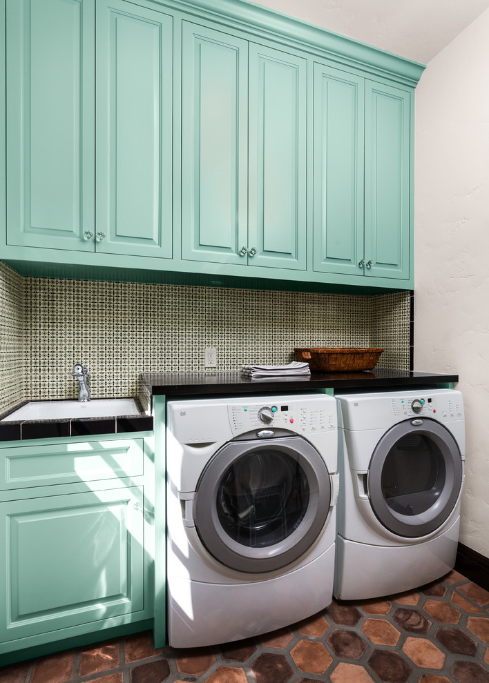 This is an example of a mid-sized mediterranean single-wall laundry room in Los Angeles with raised-panel cabinets, blue cabinets, white walls, terra-cotta floors, a side-by-side washer and dryer, brown floor, quartz benchtops and black benchtop.