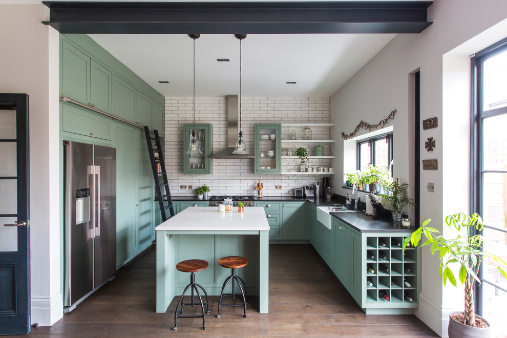 This is an example of a mid-sized contemporary u-shaped open plan kitchen in London with recessed-panel cabinets, green cabinets, medium hardwood floors, with island, brown floor, a farmhouse sink, subway tile splashback, stainless steel appliances, white splashback, granite benchtops and black benchtop.