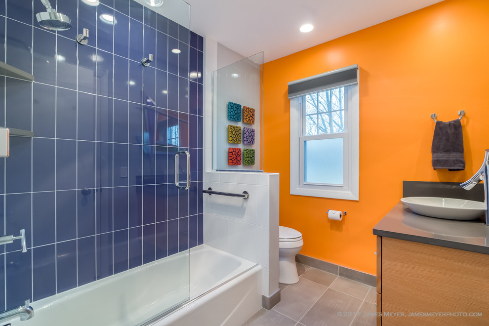 Photo of a mid-sized contemporary kids bathroom in Milwaukee with flat-panel cabinets, medium wood cabinets, an alcove tub, an alcove shower, a two-piece toilet, white tile, ceramic tile, orange walls, porcelain floors, a vessel sink, engineered quartz benchtops, grey floor, a hinged shower door and grey benchtops.