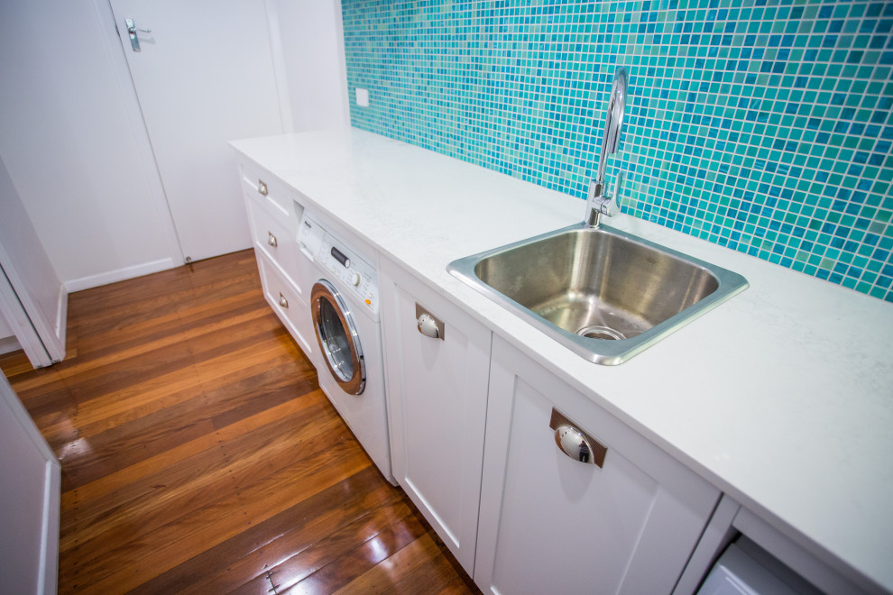 Photo of a mid-sized beach style single-wall dedicated laundry room in Brisbane with a drop-in sink, shaker cabinets, white cabinets, quartz benchtops, blue splashback, mosaic tile splashback, white walls, dark hardwood floors, a side-by-side washer and dryer, brown floor and white benchtop.