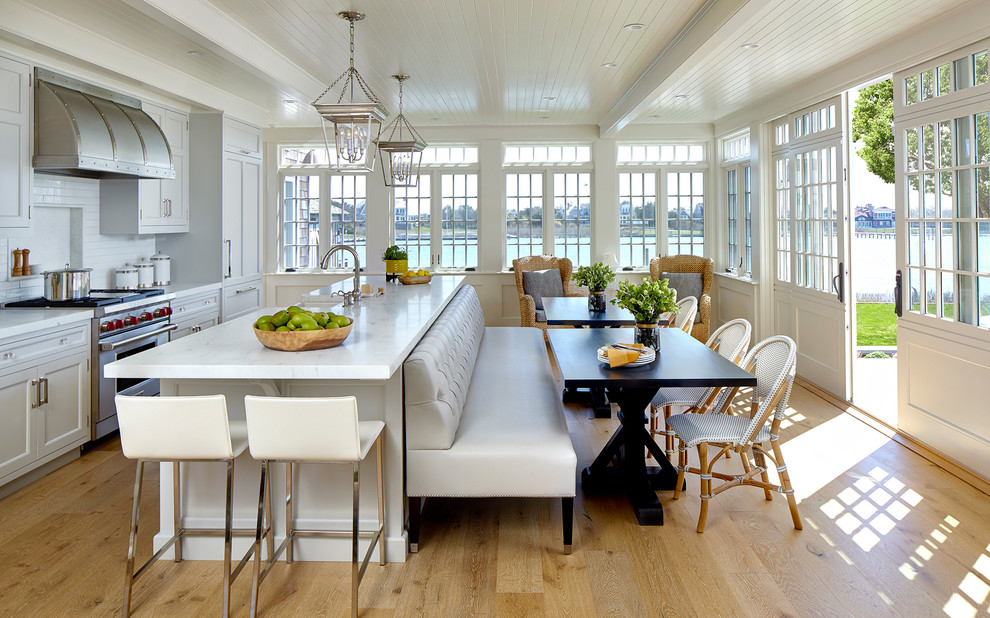 Photo of a beach style eat-in kitchen in New York with recessed-panel cabinets, grey cabinets, white splashback, subway tile splashback, stainless steel appliances, light hardwood floors, with island and white benchtop.