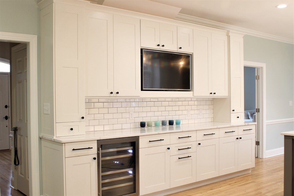 Large transitional galley light wood floor eat-in kitchen photo with a farmhouse sink, shaker cabinets, white cabinets, quartz countertops, white backsplash, ceramic backsplash, stainless steel appliances, an island and white countertops
