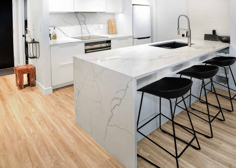 Mid-sized modern galley open plan kitchen in Vancouver with an undermount sink, flat-panel cabinets, white cabinets, onyx benchtops, white splashback, marble splashback, white appliances, light hardwood floors and a peninsula.