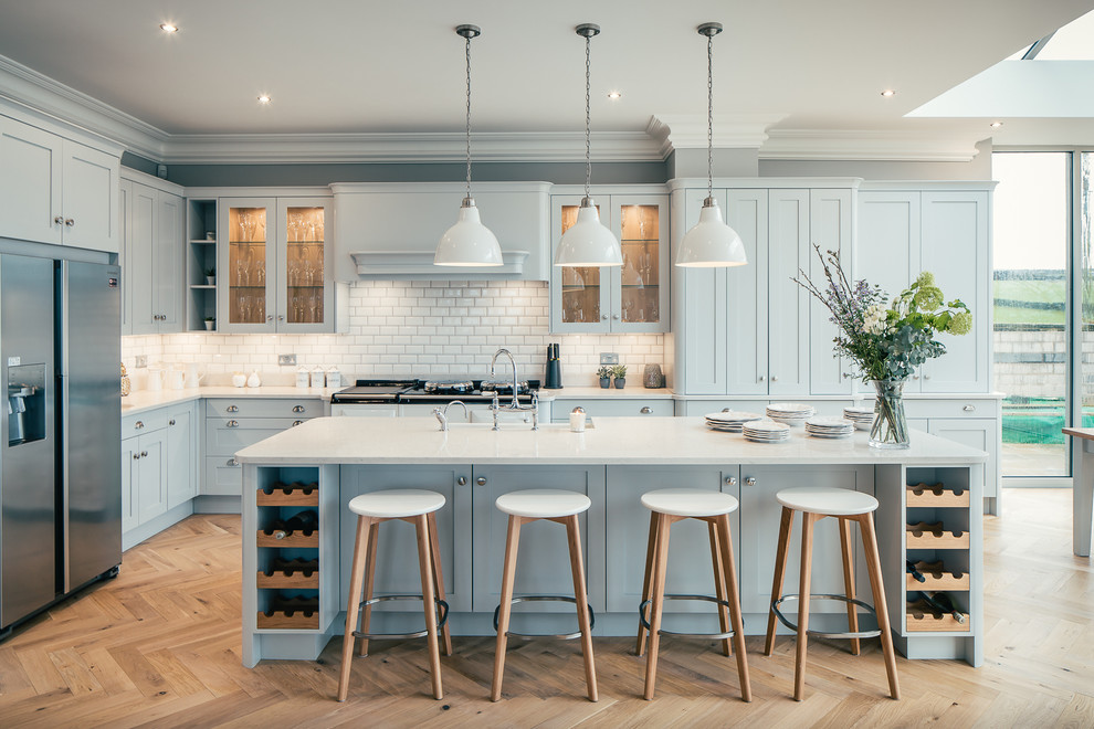 Design ideas for a large transitional l-shaped kitchen in Hertfordshire with blue cabinets, granite benchtops, white splashback, stainless steel appliances, light hardwood floors, with island, beige floor, glass-front cabinets and subway tile splashback.