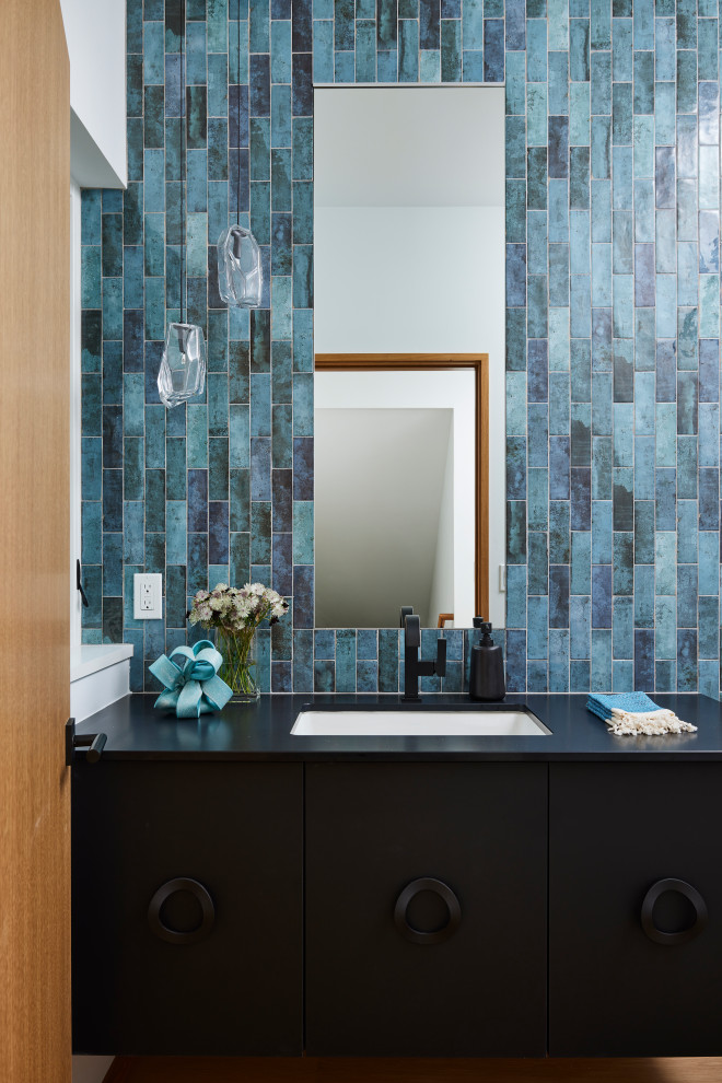 Example of a trendy blue tile medium tone wood floor and brown floor powder room design in Minneapolis with flat-panel cabinets, black cabinets, an undermount sink and black countertops