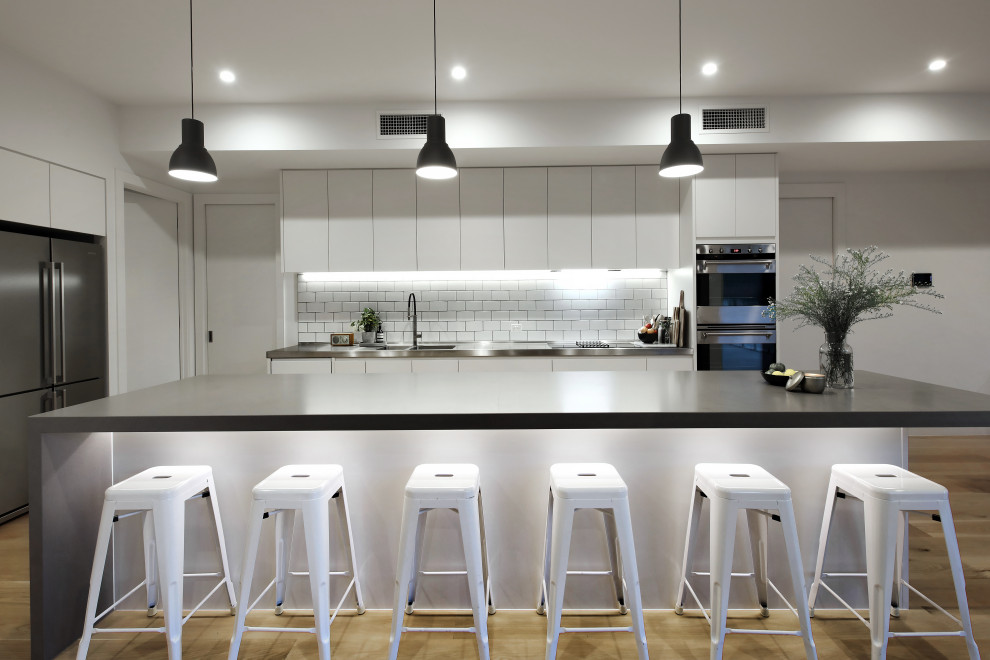 Photo of a contemporary l-shaped kitchen in Melbourne with an integrated sink, flat-panel cabinets, white cabinets, white splashback, stainless steel appliances, light hardwood floors, with island, beige floor and black benchtop.