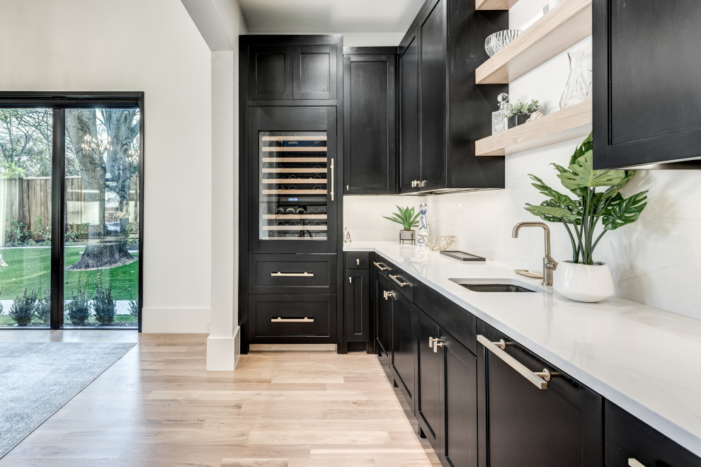 Design ideas for a mid-sized transitional l-shaped wet bar in Dallas with an undermount sink, recessed-panel cabinets, black cabinets, quartz benchtops, white splashback, marble splashback, light hardwood floors, brown floor and white benchtop.