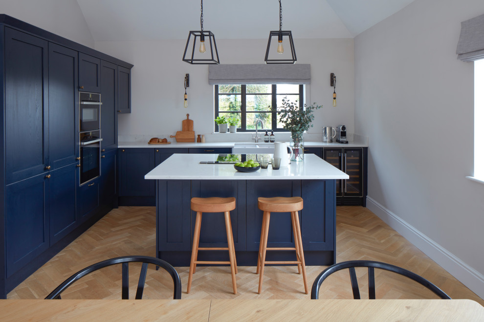 Country Wohnküche in L-Form mit Landhausspüle, Schrankfronten im Shaker-Stil, blauen Schränken, schwarzen Elektrogeräten, hellem Holzboden, Kücheninsel, beigem Boden, weißer Arbeitsplatte und gewölbter Decke in Sussex