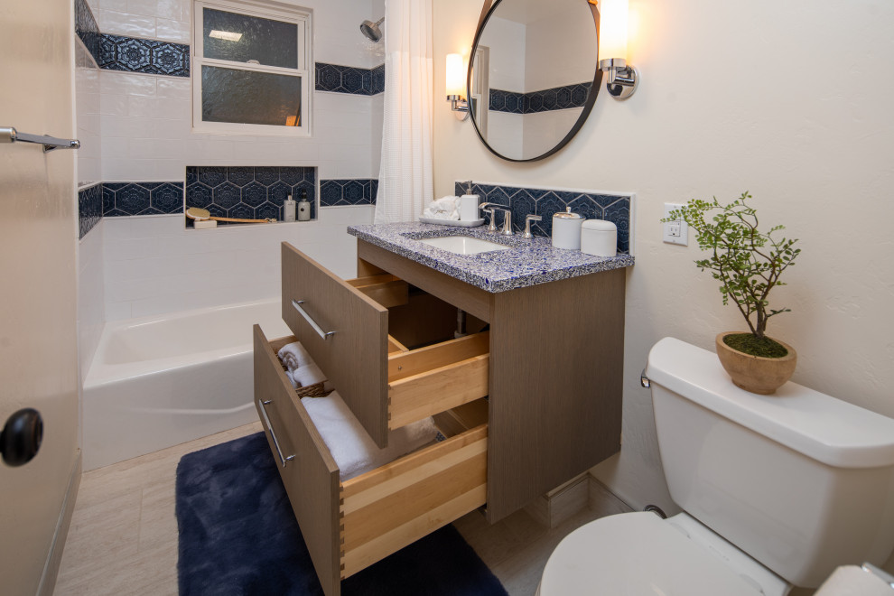 Klassisk inredning av ett litet blå blått badrum, med släta luckor, bruna skåp, ett badkar i en alkov, en dusch/badkar-kombination, en toalettstol med separat cisternkåpa, blå kakel, keramikplattor, beige väggar, klinkergolv i porslin, ett undermonterad handfat, bänkskiva i återvunnet glas, beiget golv och dusch med duschdraperi