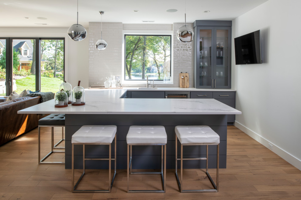 This is an example of a contemporary u-shaped seated home bar in Minneapolis with an undermount sink, glass-front cabinets, grey cabinets, white splashback, light hardwood floors and white benchtop.