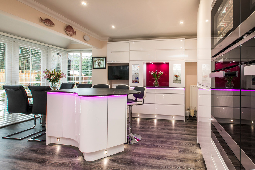 Inspiration for a large modern u-shaped kitchen in Hampshire with an integrated sink, flat-panel cabinets, white cabinets, pink splashback, glass sheet splashback, stainless steel appliances, with island, brown floor and black benchtop.
