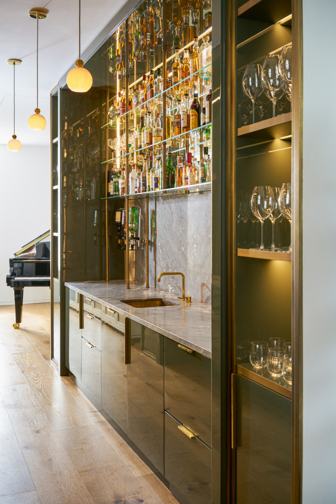 Inspiration for a large contemporary galley home bar in London with a drop-in sink, flat-panel cabinets, green cabinets, marble benchtops, marble splashback and light hardwood floors.