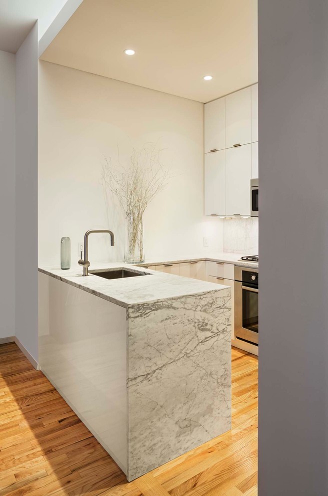This is an example of a small modern u-shaped eat-in kitchen in New York with an undermount sink, flat-panel cabinets, white cabinets, marble benchtops, white splashback, marble splashback, stainless steel appliances, light hardwood floors, a peninsula and yellow floor.