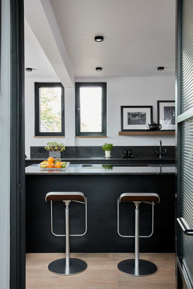 Photo of a small contemporary grey and black l-shaped open plan kitchen in London with a submerged sink, flat-panel cabinets, black cabinets, quartz worktops, grey splashback, engineered quartz splashback, stainless steel appliances, light hardwood flooring, an island, brown floors and grey worktops.