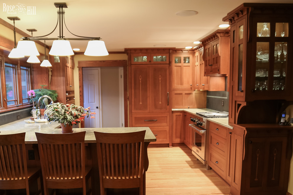 This is an example of a mid-sized arts and crafts l-shaped eat-in kitchen in Indianapolis with a farmhouse sink, recessed-panel cabinets, medium wood cabinets, granite benchtops, grey splashback, stone tile splashback, panelled appliances and light hardwood floors.