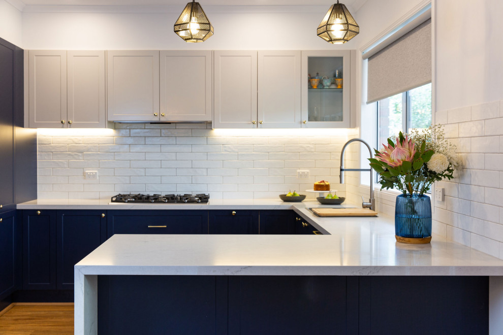 Design ideas for a large modern u-shaped kitchen pantry in Melbourne with a double-bowl sink, shaker cabinets, blue cabinets, solid surface benchtops, white splashback, subway tile splashback, stainless steel appliances, medium hardwood floors, with island and white benchtop.