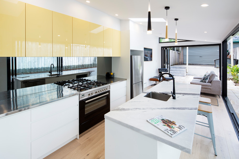 Photo of a contemporary galley kitchen in Newcastle - Maitland with an undermount sink, flat-panel cabinets, yellow cabinets, stainless steel benchtops, black splashback, glass sheet splashback, stainless steel appliances, medium hardwood floors, with island, brown floor and grey benchtop.