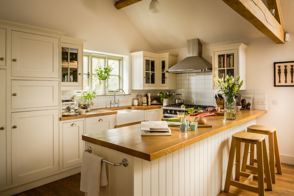 Photo of a mid-sized country u-shaped open plan kitchen in Wiltshire with a farmhouse sink, shaker cabinets, white cabinets, wood benchtops, white splashback, ceramic splashback, black appliances, medium hardwood floors and beige floor.