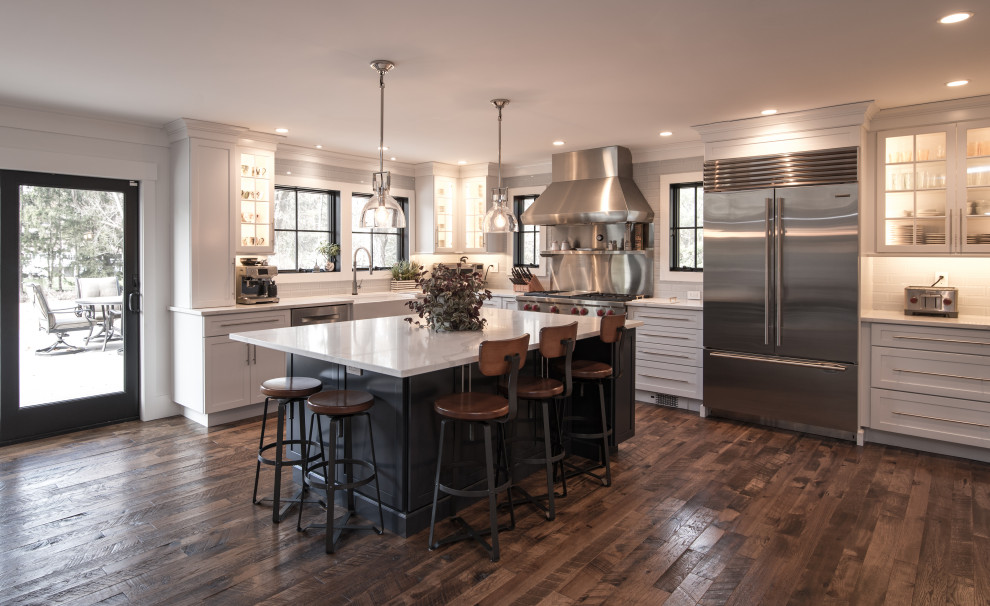 Photo of a large modern l-shaped eat-in kitchen in New York with shaker cabinets, white cabinets, white splashback, stainless steel appliances, dark hardwood floors, with island, brown floor and white benchtop.
