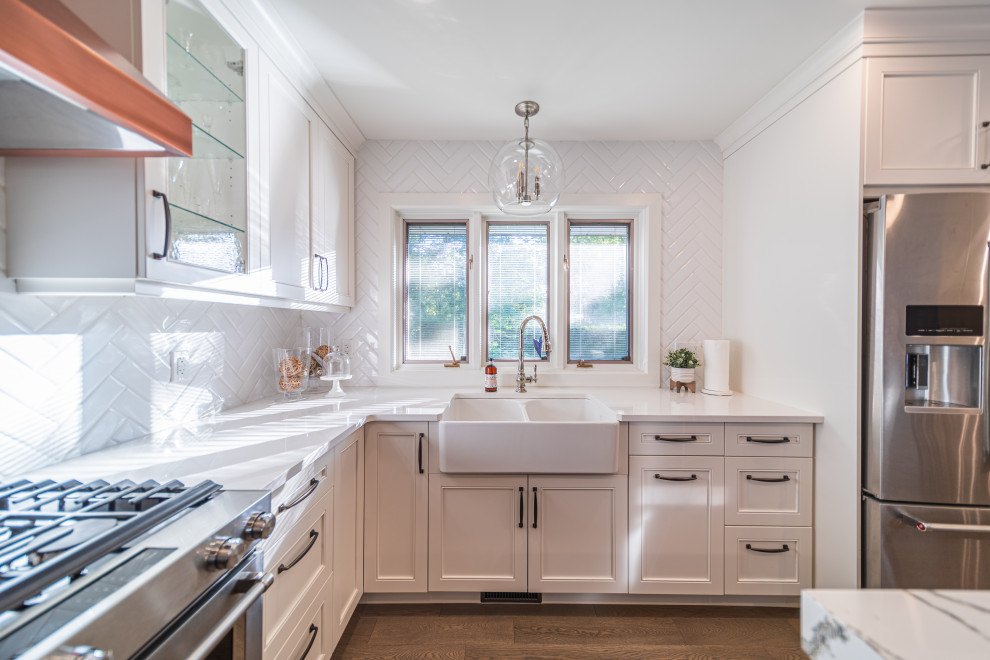 Photo of a mid-sized transitional l-shaped eat-in kitchen in Toronto with a farmhouse sink, recessed-panel cabinets, white cabinets, quartzite benchtops, white splashback, subway tile splashback, stainless steel appliances, medium hardwood floors, with island, brown floor and white benchtop.