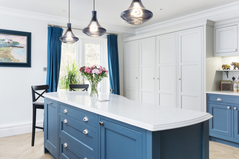 This is an example of a large classic single-wall open plan kitchen in Cardiff with a belfast sink, shaker cabinets, blue cabinets, engineered stone countertops, white splashback, black appliances, light hardwood flooring, an island, brown floors and white worktops.