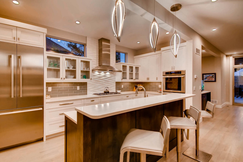 Photo of a contemporary l-shaped open plan kitchen in Denver with shaker cabinets, white cabinets, engineered stone countertops, grey splashback, metro tiled splashback, stainless steel appliances, light hardwood flooring, an island and white worktops.