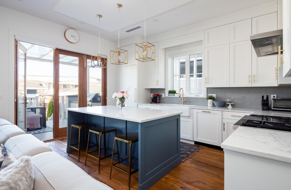 Example of a transitional medium tone wood floor and brown floor kitchen design in Vancouver with a farmhouse sink, shaker cabinets, white cabinets, quartz countertops, mosaic tile backsplash, stainless steel appliances, an island and white countertops