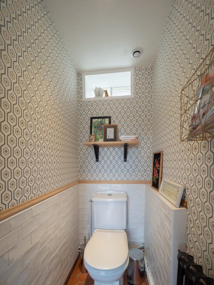 Foto på ett litet lantligt toalett, med en toalettstol med hel cisternkåpa, vit kakel, tunnelbanekakel, klinkergolv i terrakotta och brunt golv