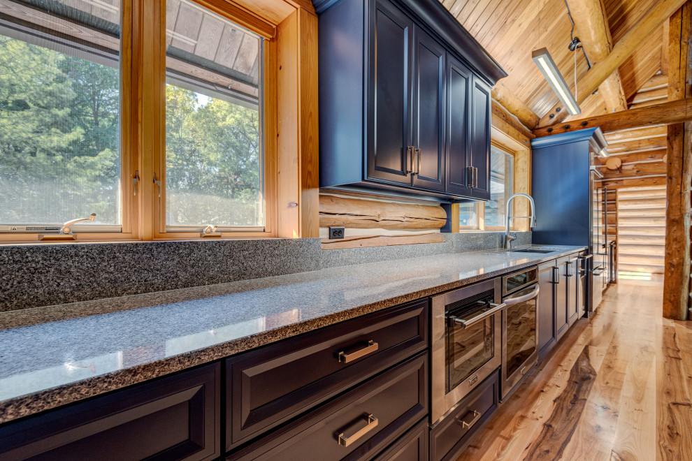 Cette photo montre une cuisine américaine parallèle chic avec un évier encastré, un placard à porte shaker, des portes de placard bleues, un plan de travail en quartz, une crédence en bois, un électroménager en acier inoxydable, un sol en bois brun, îlot et un plan de travail gris.