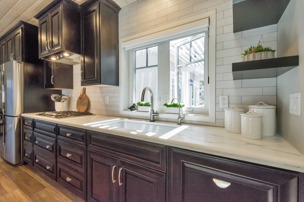 Photo of a large transitional single-wall open plan kitchen in Phoenix with a farmhouse sink, raised-panel cabinets, black cabinets, quartzite benchtops, white splashback, porcelain splashback, stainless steel appliances, porcelain floors, with island and brown floor.