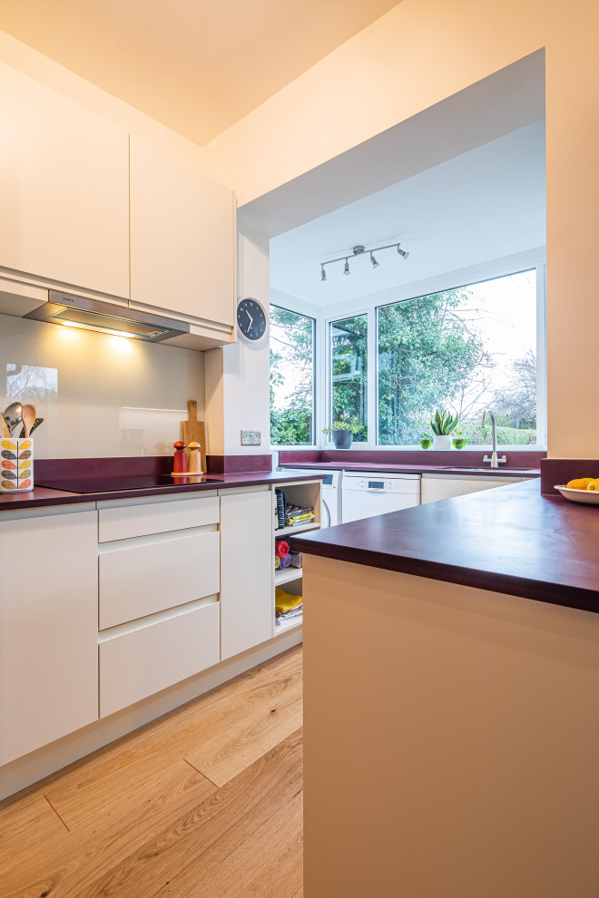 Small contemporary u-shaped eat-in kitchen in Other with flat-panel cabinets, white cabinets, white splashback, glass sheet splashback, stainless steel appliances, no island, brown floor and red benchtop.