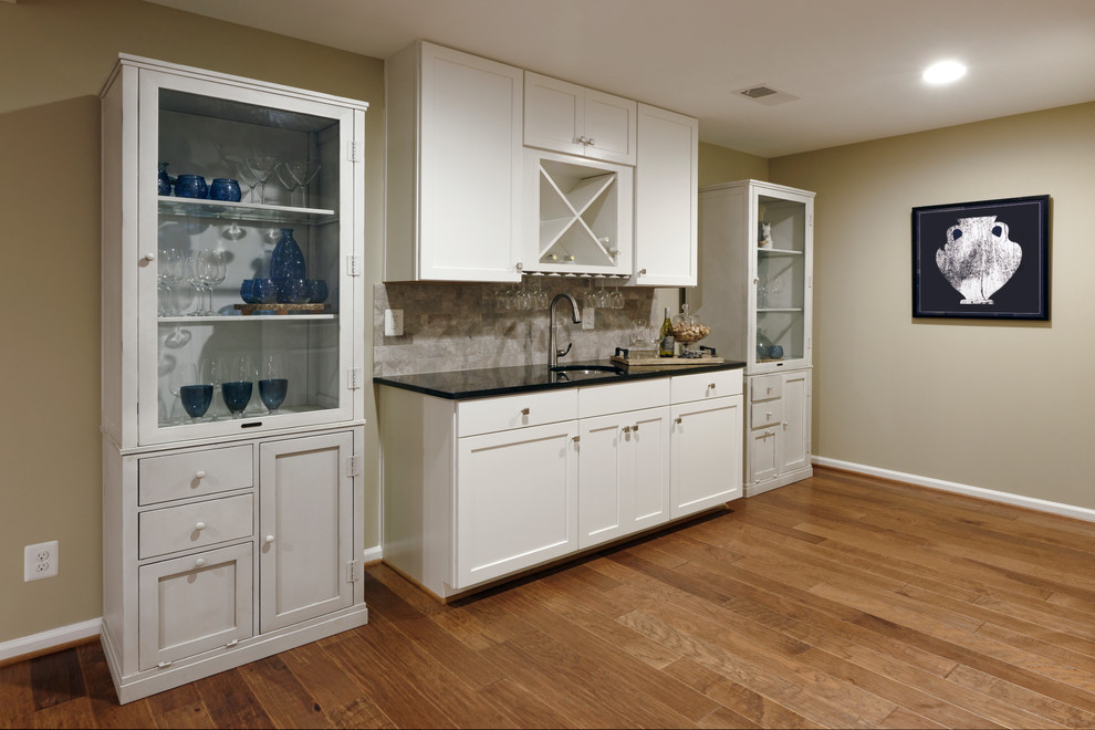 This is an example of a mid-sized transitional single-wall wet bar in DC Metro with an undermount sink, shaker cabinets, white cabinets, solid surface benchtops, multi-coloured splashback, subway tile splashback, vinyl floors and brown floor.