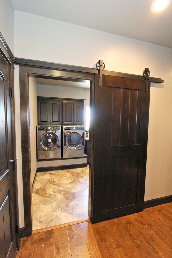 Photo of an expansive traditional l-shaped dedicated laundry room in Boise with an undermount sink, raised-panel cabinets, dark wood cabinets, granite benchtops, beige walls, travertine floors and a side-by-side washer and dryer.
