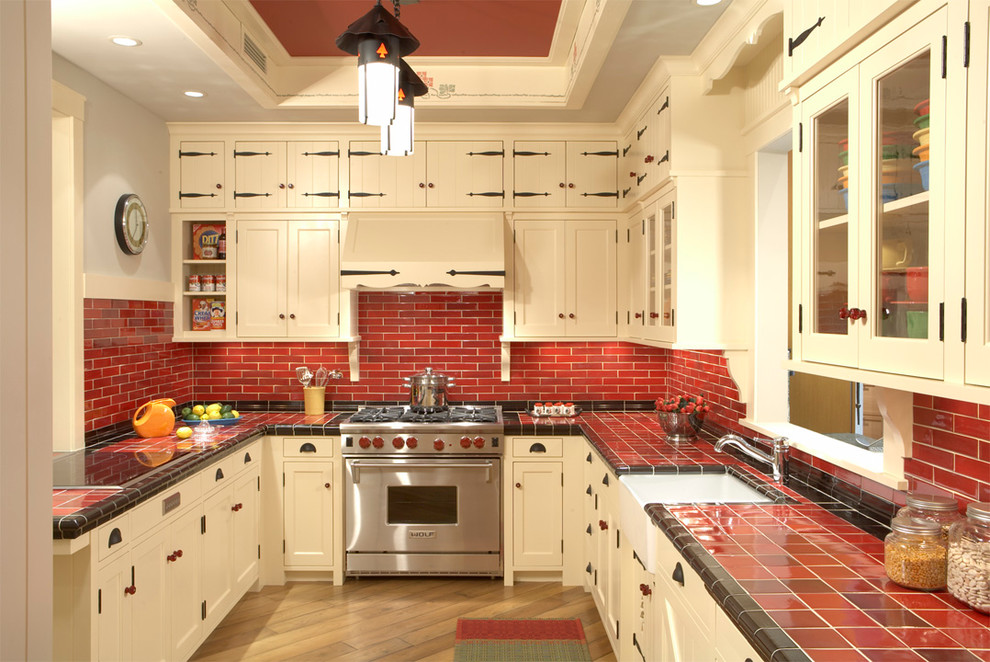 This is an example of a country u-shaped separate kitchen in Minneapolis with stainless steel appliances, red splashback, tile benchtops, beige cabinets, recessed-panel cabinets, a farmhouse sink, ceramic splashback, medium hardwood floors, a peninsula and red benchtop.