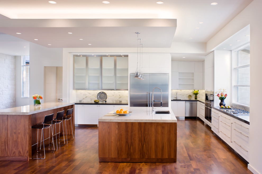 Inspiration for a large contemporary galley open plan kitchen in Austin with an integrated sink, glass-front cabinets, stainless steel cabinets, white splashback, stone tile splashback, stainless steel appliances, dark hardwood floors, multiple islands, marble benchtops and brown floor.