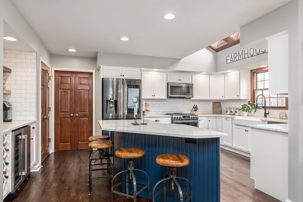 Photo of a large country u-shaped eat-in kitchen in Columbus with shaker cabinets, quartz benchtops, white splashback, with island, a farmhouse sink, white cabinets, subway tile splashback, stainless steel appliances, medium hardwood floors, brown floor and white benchtop.